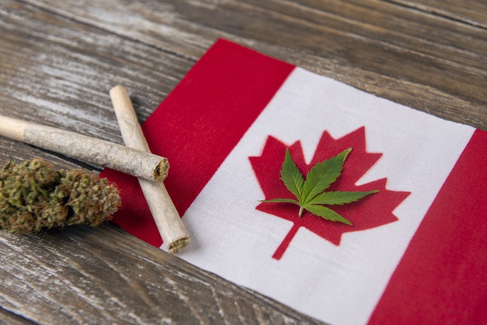 A cannabis leaf laid within the outline of the red maple leaf in Canada's flag, with two rolled joints and a cannabis bud next to the flag.