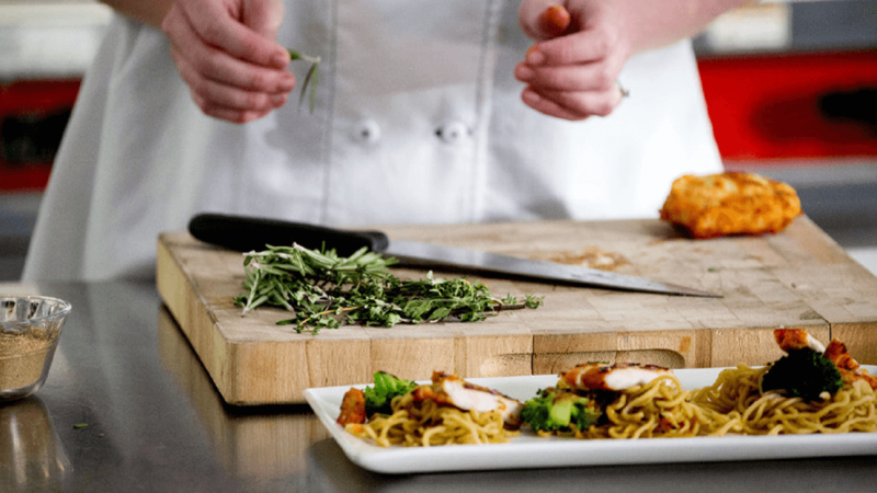 Cutting board with chopped vegetables on it in front of a plate of food, with chef standing behind.