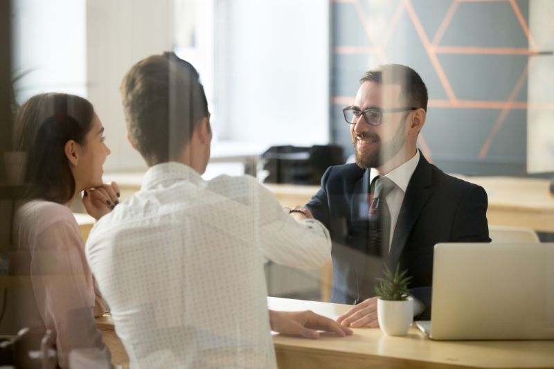 A banker shakes hands across his desk with a young couple.