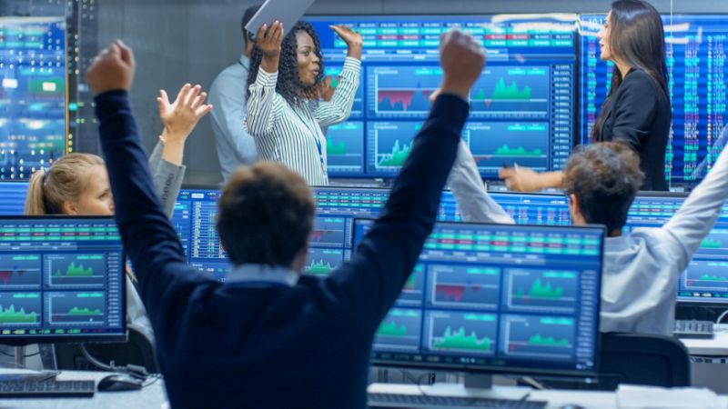 Traders celebrating in front of colorful monitors displaying stock market data.