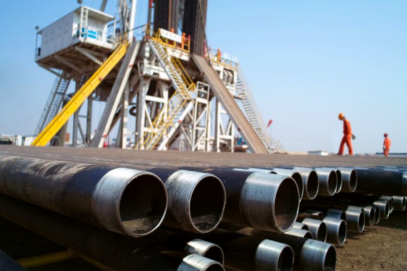 Rig and pipe with two workers wearing orange clothes.