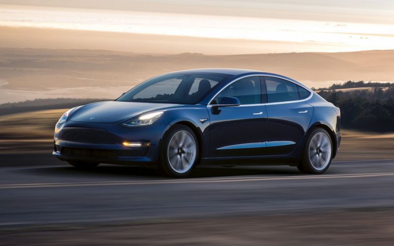 Dark blue Model 3 sedan on a road in front of a picturesque landscape.