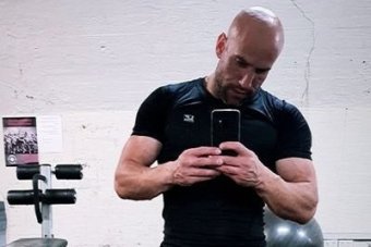 Stavro D'Amore wearing a tight black shirt with muscles exposed, wearing grey pants, standing in a gym taking a photo of himself