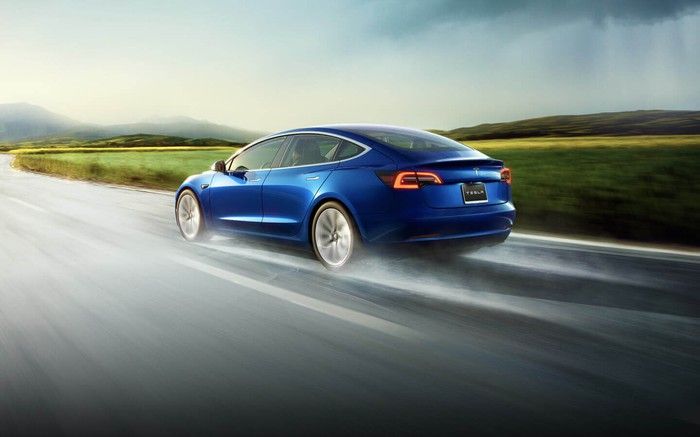 Blue Tesla Model 3 on a wet road on a cloudy day, driving through a grassy landscape.