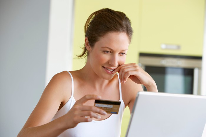 A smiling woman holding a credit card in her right hand while looking at an open laptop. 